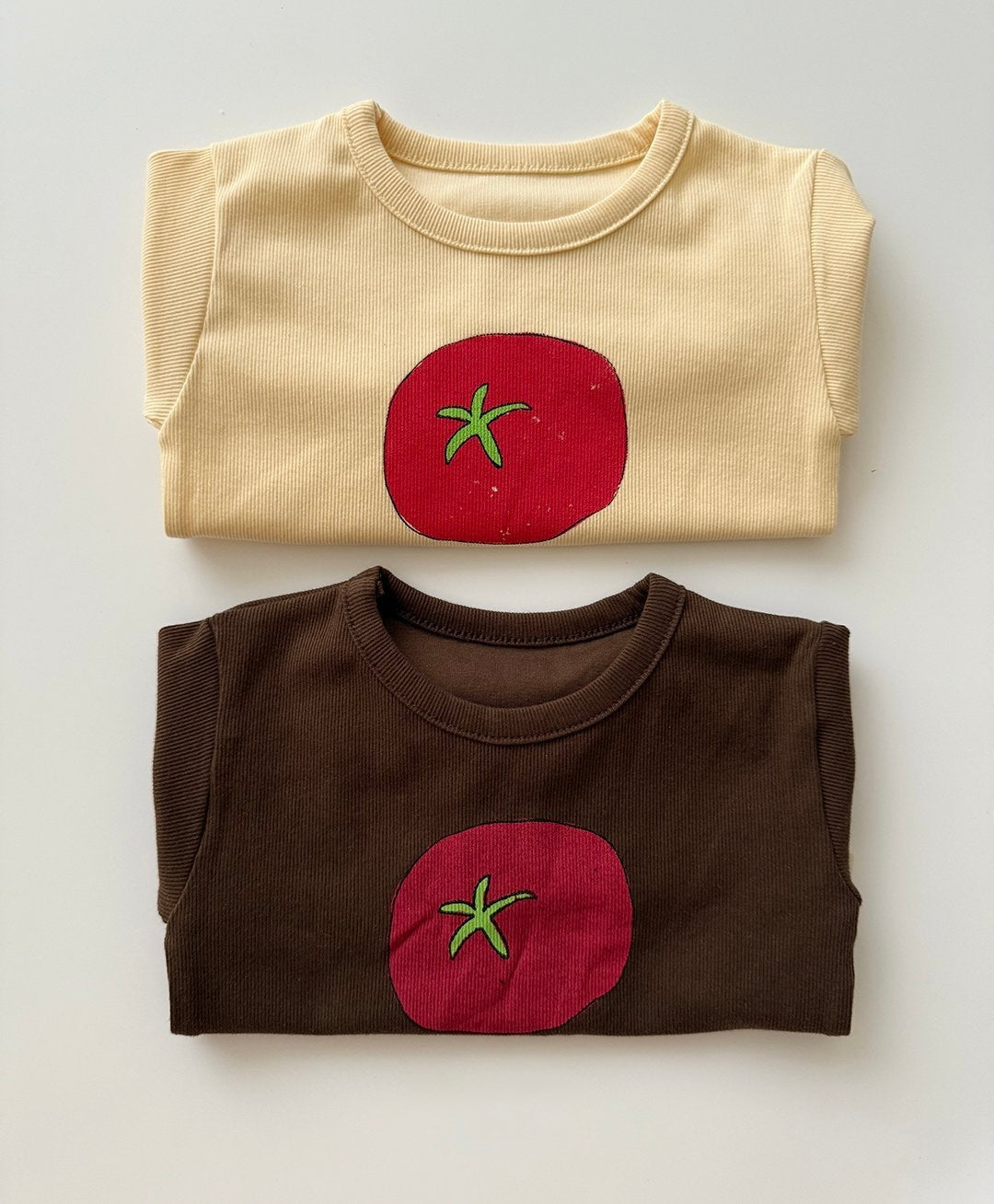 Two folded shirts, one beige and one brown, with tomato designs for toddlers and baby